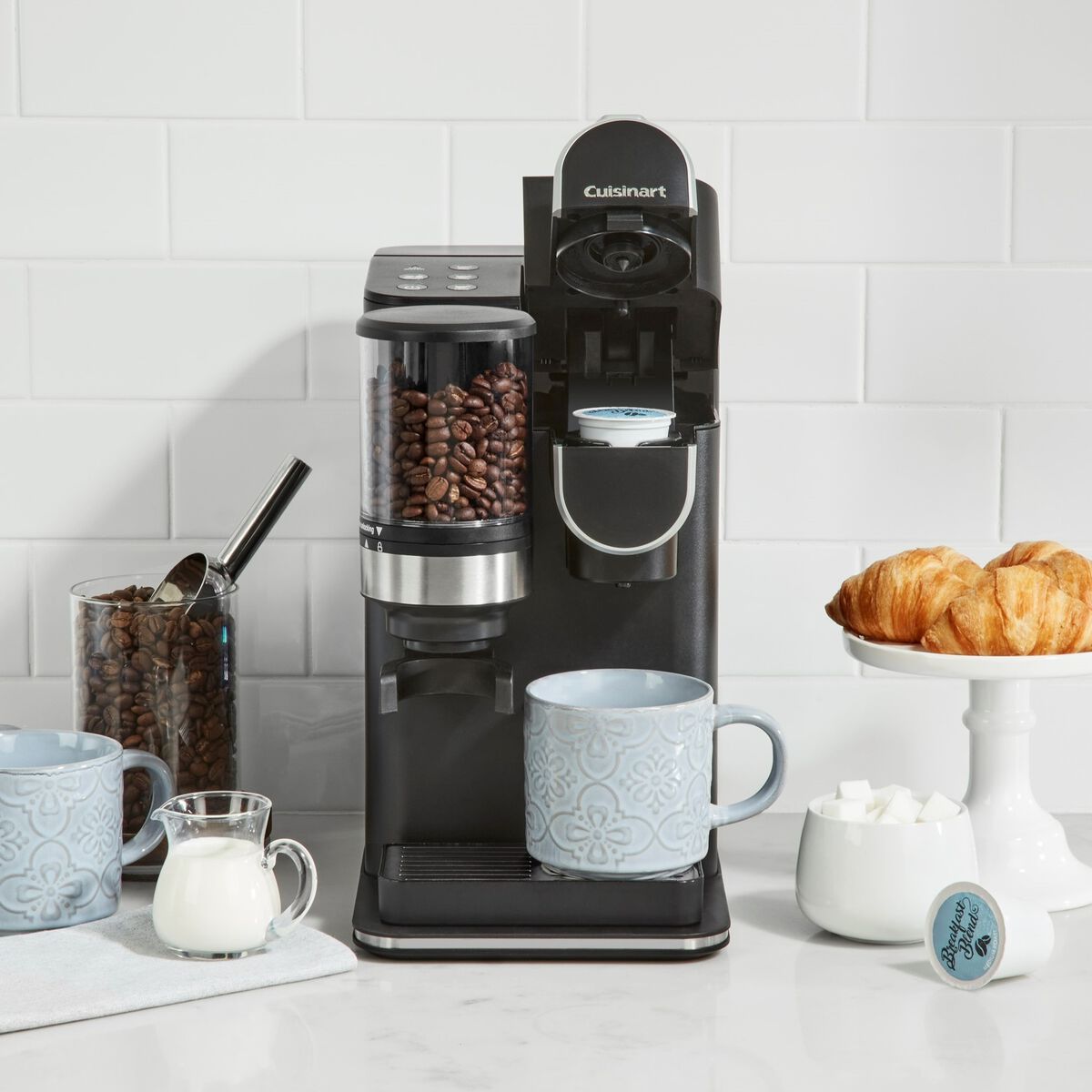 Cuisinart DGB-2 Grind & Brew Single-Serve coffee maker in matte black on a kitchen counter — top-loaded whole-bean hopper, pod tray loaded with a K-Cup, cup underneath the spout, extra beans and pastries staged around it to show the whole-bean-or-pod flex.
