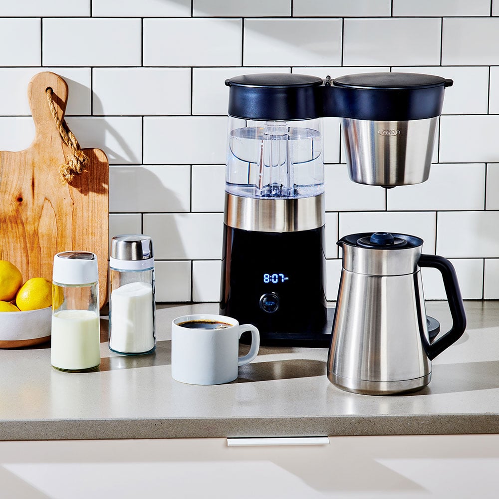 OXO Brew 9-Cup Barista Brain coffee maker on a countertop with subway-tile backsplash — single-dial UX reading '8:07 AM', double-wall stainless thermal carafe beside the machine, small cup of brewed coffee in front.